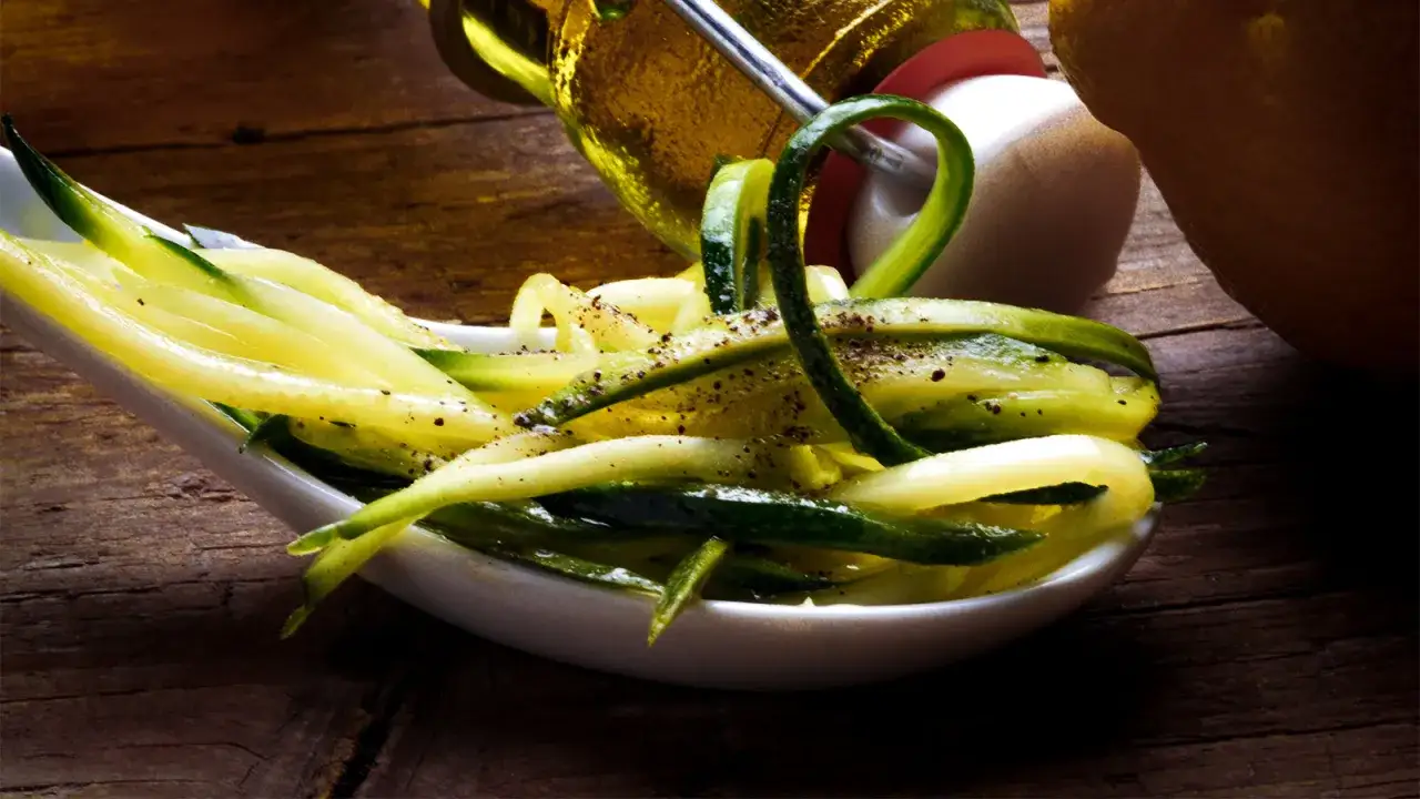 Zucchine marinate fresche, condite con pepe nero, servite su un cucchiaio bianco, con una bottiglia d'olio e un limone sullo sfondo.