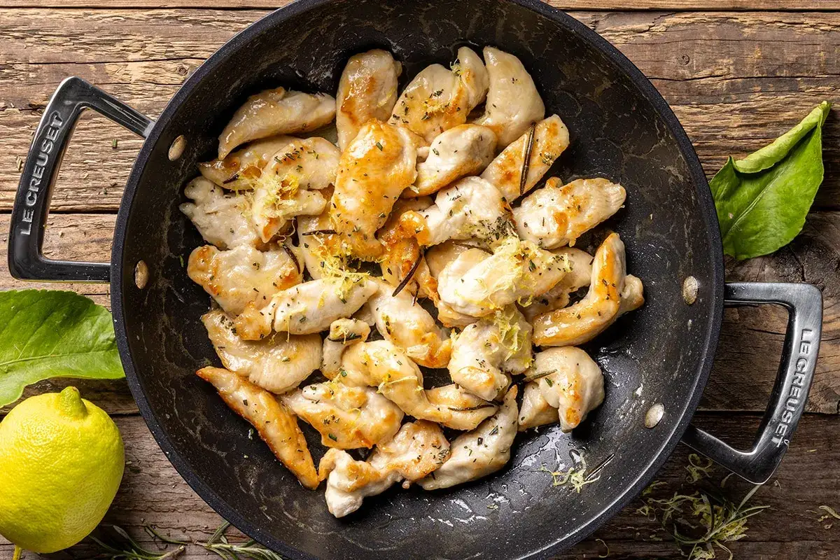 Bocconcini di pollo dorati e succulenti, conditi con erbe aromatiche e scorza di limone, in una padella Le Creuset su un tavolo di legno.