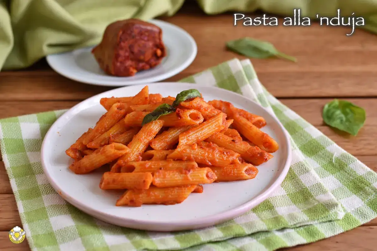 Piatto di penne condite con un sugo rosso piccante alla 'nduja, guarnito con basilico. Sullo sfondo, un pezzo di 'nduja.