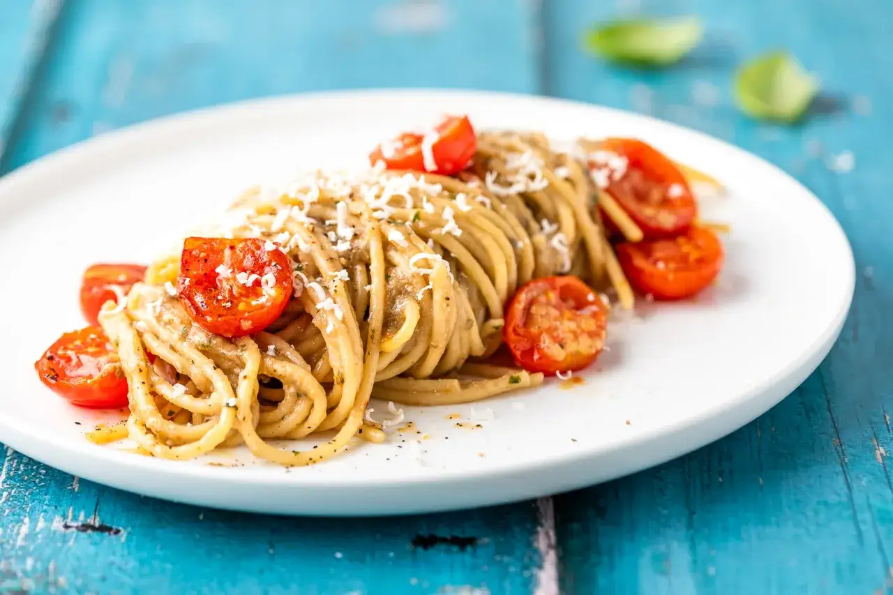 Spaghetti con sugo di melanzane, pomodorini confit e parmigiano grattugiato su un piatto bianco.