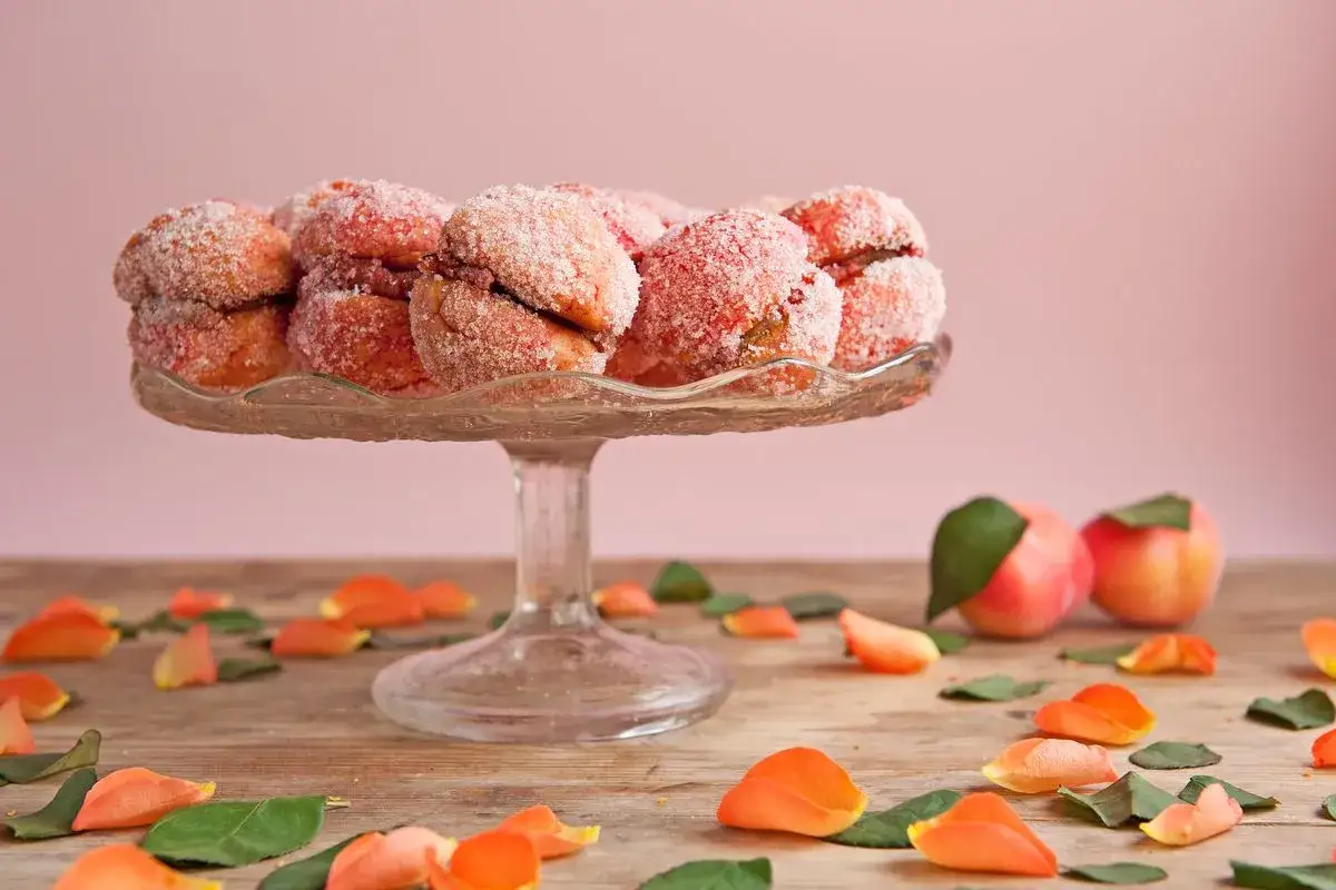 Dolci pesche ricoperte di zucchero su un'alzata di vetro, con petali e foglie sparsi sul tavolo.