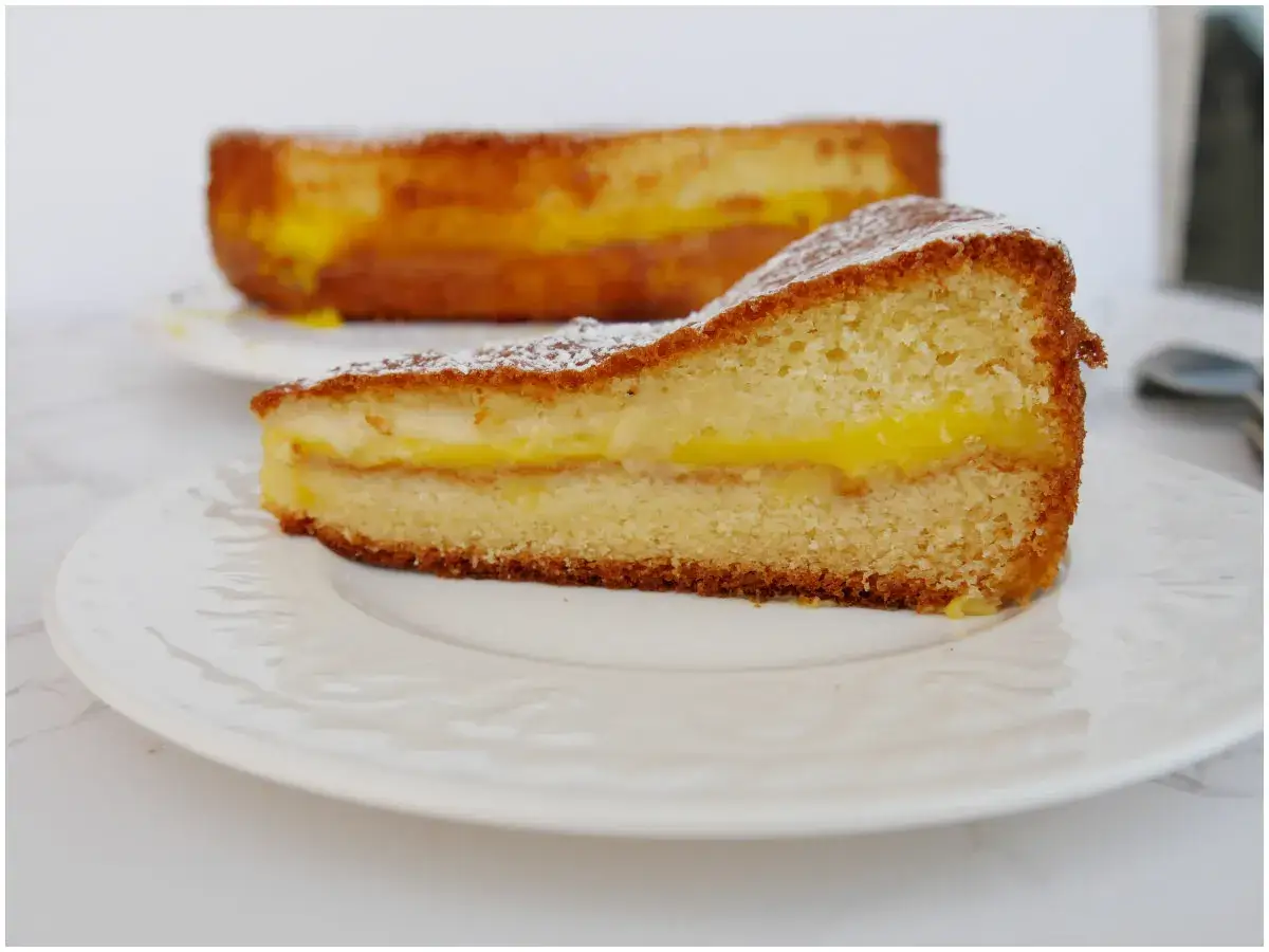 Una fetta di torta con crema pasticcera, soffice e dorata, cosparsa di zucchero a velo. Sullo sfondo, un'altra torta simile.