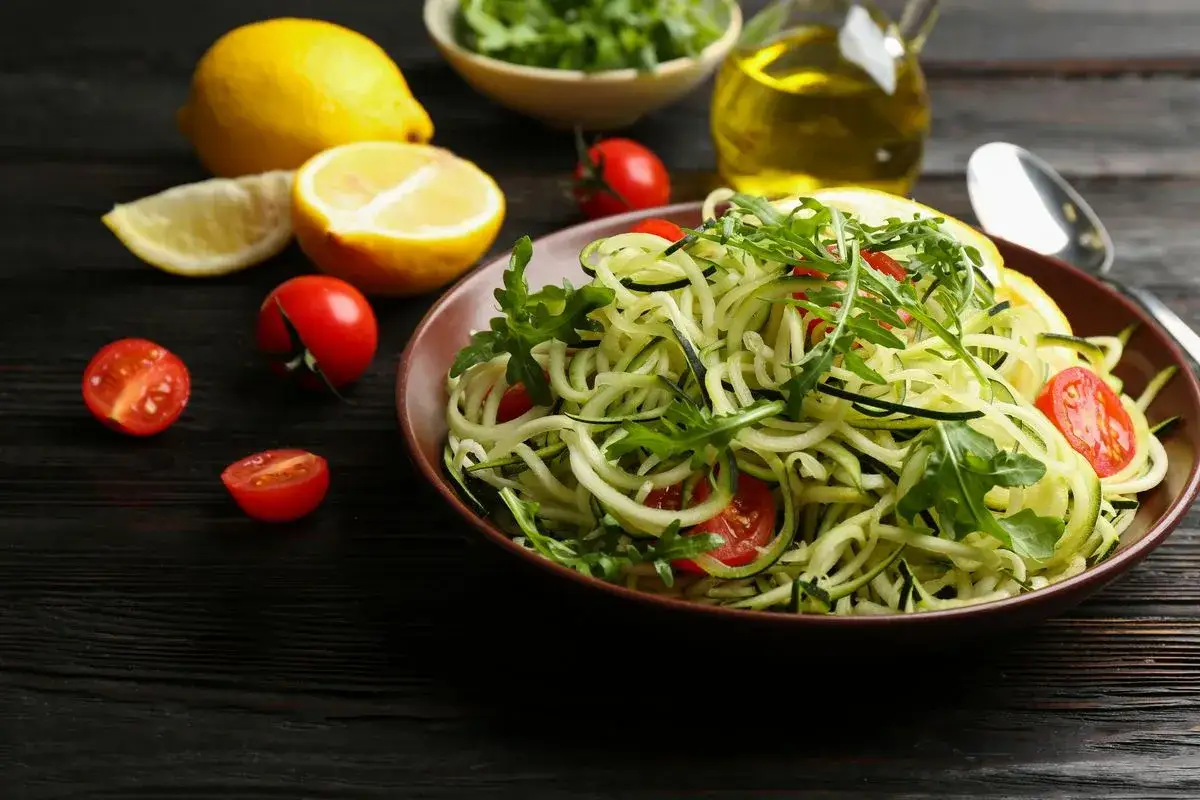 Un piatto di spaghetti di zucchine freschi, guarniti con pomodorini e rucola, pronti per essere conditi con limone e olio d'oliva.