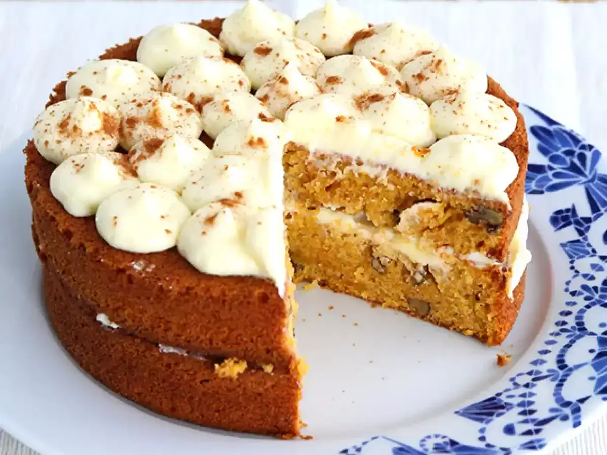 Torta carote e noci soffice, con glassa bianca e spolverata di cannella. Una fetta è stata tolta, rivelando strati golosi.