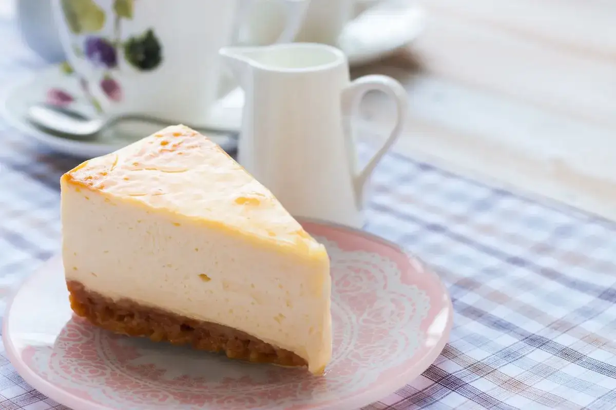 Una fetta di cheesecake con mascarpone, cremosa e invitante, su un piattino rosa. Sullo sfondo, una tazza da t&egrave; e una lattiera.