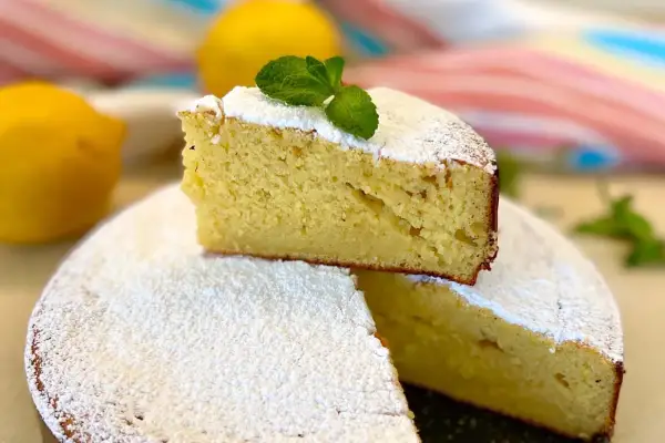 Una fetta di torta allo yogurt greco, soffice e spolverata di zucchero a velo, decorata con menta fresca. Due limoni sullo sfondo.