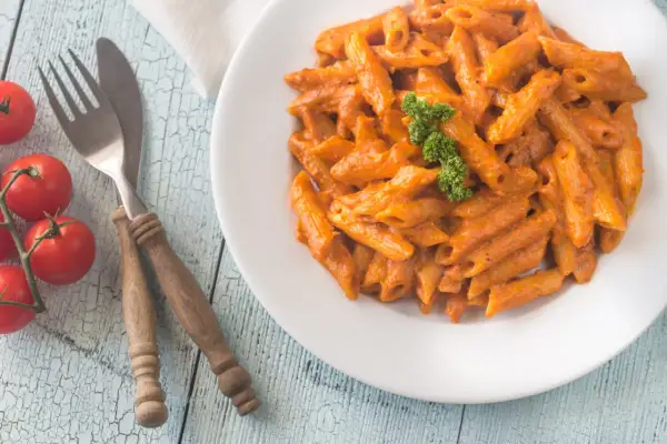 Piatto di penne alla vodka cremose, guarnite con prezzemolo, su un tavolo di legno azzurro. Pomodorini freschi e posate completano la scena.