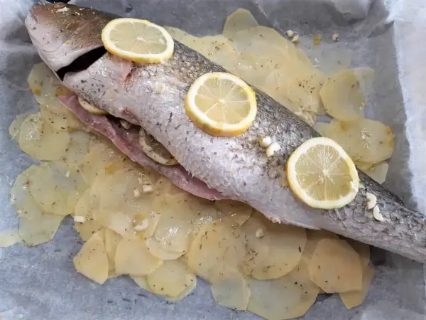 Ombrina al forno pronta per essere infornata, adagiata su patate affettate e guarnita con fette di limone e aglio.