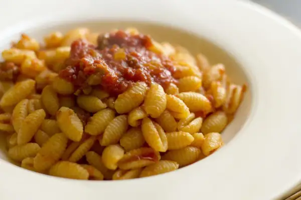 Un piatto di malloreddus alla campidanese, pasta sarda con sugo di pomodoro e salsiccia, serviti in una ciotola bianca.