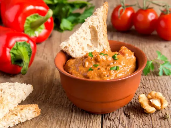 Pesto di peperoni in ciotola di terracotta, con pane tostato, noci e peperoni rossi sullo sfondo.