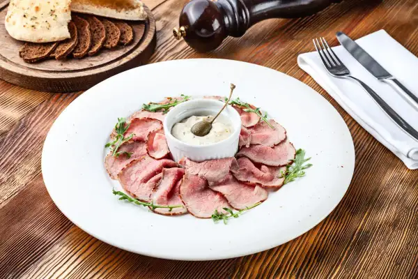 Fettine di arrosto rosa disposte a cerchio su un piatto bianco, con una ciotolina di salsa tonnata guarnita da un cappero.