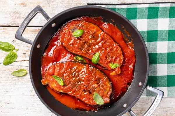 Melanzane in padella, succulente e cotte in un ricco sugo di pomodoro, guarnite con foglie di basilico fresco.