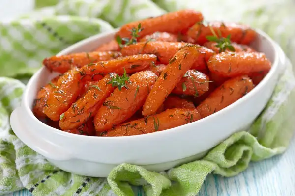 Carote glassate in padella, profumate con erbe aromatiche, servite in una ciotola bianca su un tovagliolo verde.