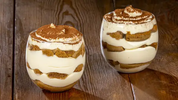 Due bicchieri di tiramisù senza uova, stratificati con crema e savoiardi, spolverati di cacao.