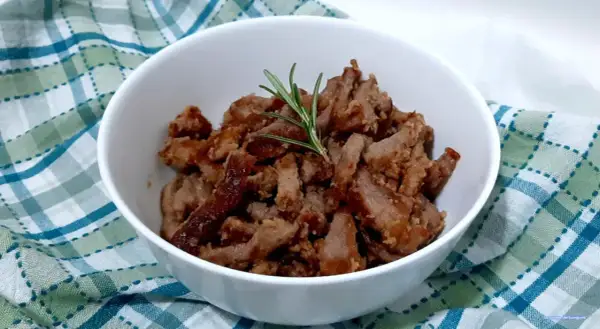 Deliziosi straccetti di vitello cotti e guarniti con un rametto di rosmarino, serviti in una ciotola bianca su un tovagliolo a quadri.