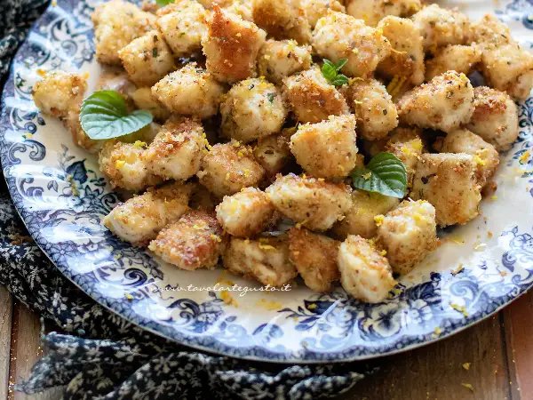 Bocconcini di pollo dorati e croccanti, guarniti con scorza di limone e foglioline di menta, serviti su un piatto decorato.