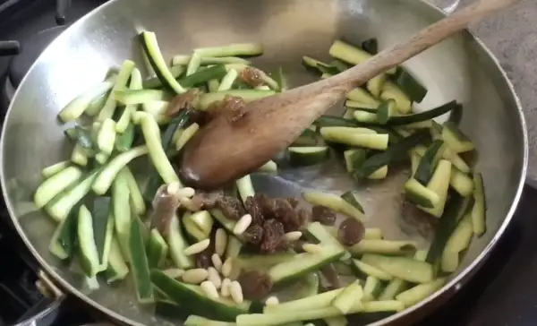 Zucchine tagliate a julienne con uvetta e pinoli in padella, pronte per essere trasformate in un delizioso piatto agrodolce.