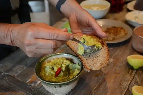 Mani che spalmano la salsa guacamole ricetta su una fetta di pane tostato. Un peperoncino rosso decora la ciotola.