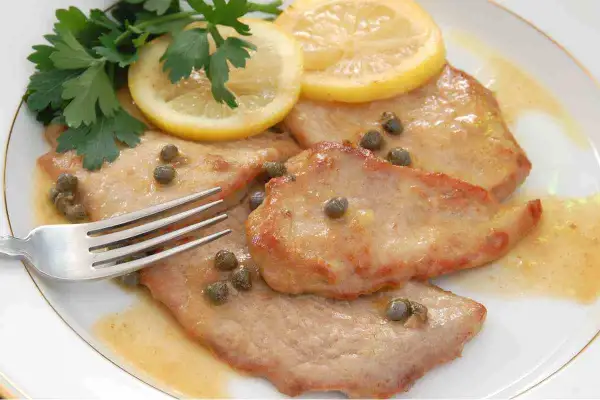 Scaloppine al limone cremose, guarnite con capperi, prezzemolo e fette di limone, pronte per essere gustate con una forchetta.