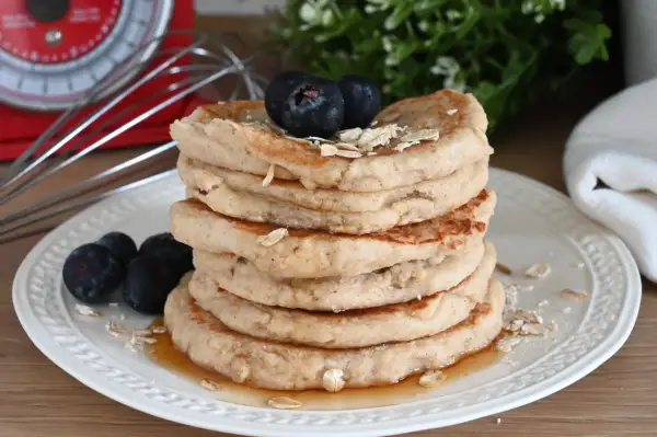 Pila di pancake con farina di avena, guarniti con mirtilli e sciroppo d'acero. Sullo sfondo, una bilancia rossa e un frullino.
