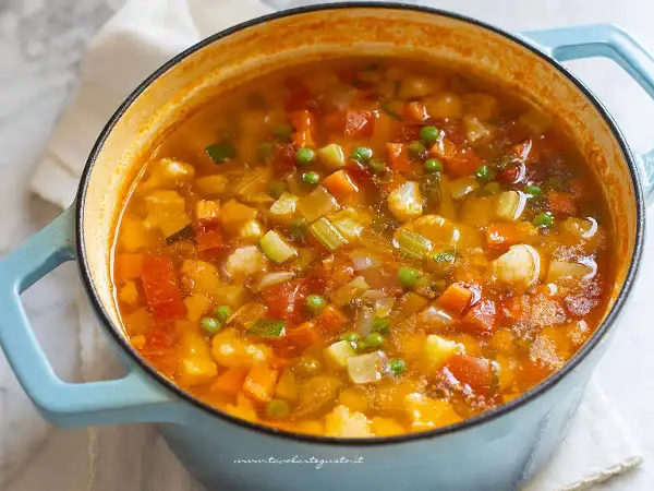 Un fumante minestrone di verdure in una casseruola azzurra, ricco di carote, zucchine, piselli e pomodoro.