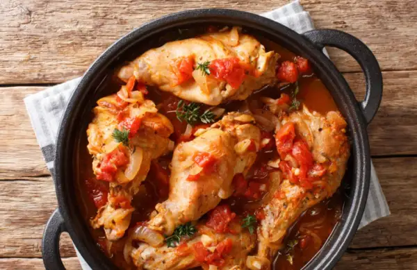 Coniglio all'ischitana in tegame, con pomodoro, cipolla e erbe aromatiche, servito su un tavolo di legno.