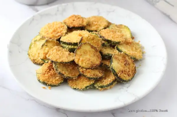 Fettine di zucchine impanate e cotte nella friggitrice ad aria, dorate e croccanti, servite su un piatto bianco.