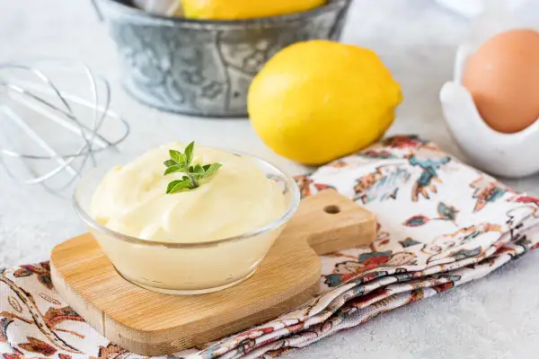 Una ciotola di cremosa maionese fatta in casa, guarnita con erbe fresche, accanto a limoni, un uovo e una frusta.